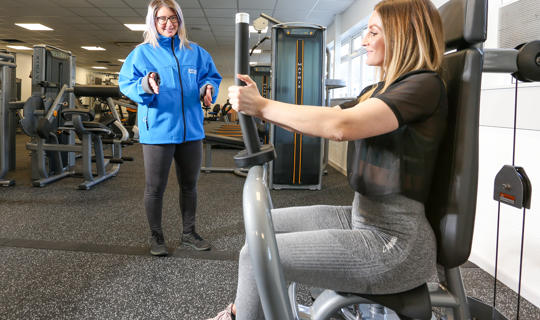 Woman standing up talking to another women who is sat on strength equipment in a gym