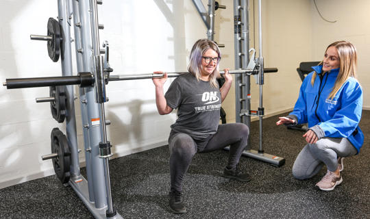 Woman lifting a weight in a gym with another woman watching her